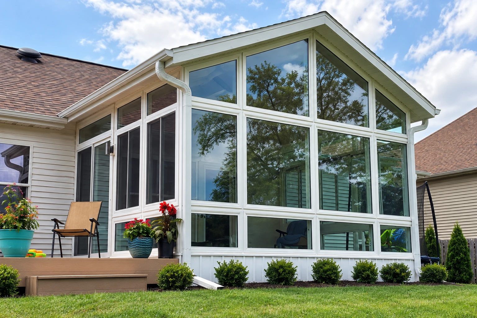 White-framed sunroom addition with tall gabled windows beside a wooden deck and landscaped lawn.