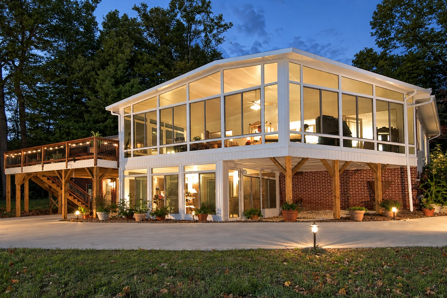 Two-story sunroom addition with expansive white-framed windows glows beside a raised wooden deck at dusk.