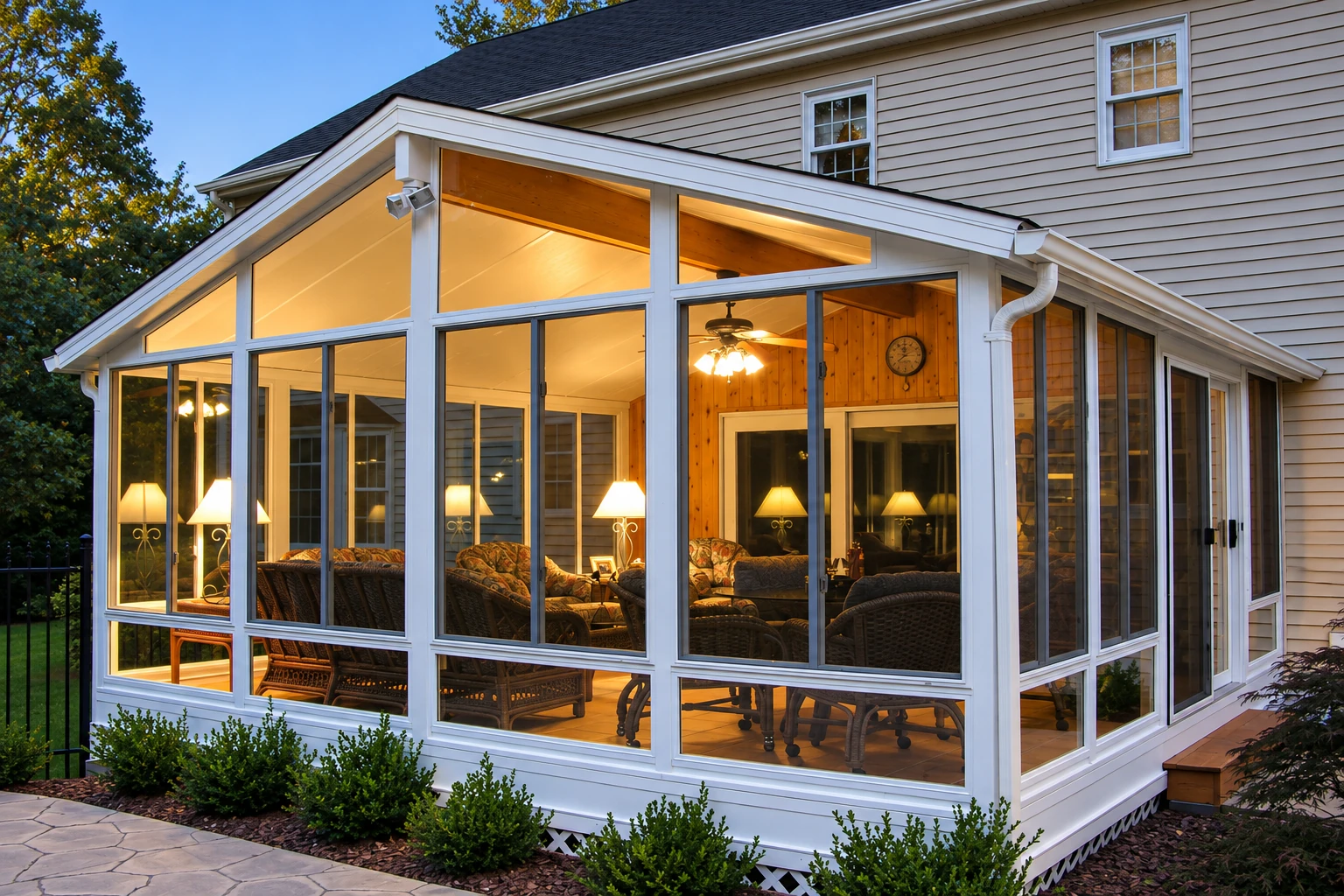 White vinyl sunroom with a peaked roof and large windows attached to a house, warmly lit inside.